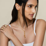 Woman wearing gold jewelry including earrings, necklace, and ring on a neutral background