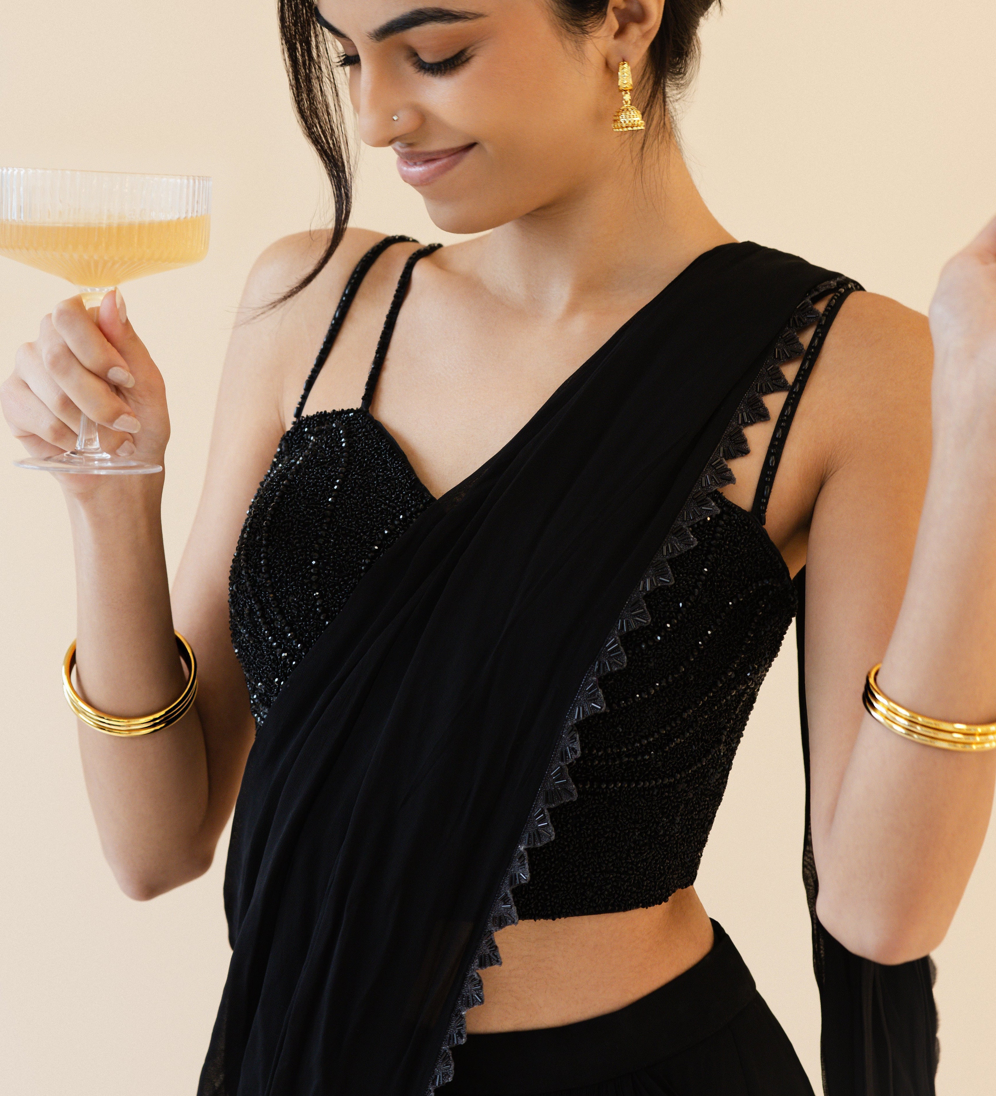 Woman in a black saree holding a glass of champagne against a beige background