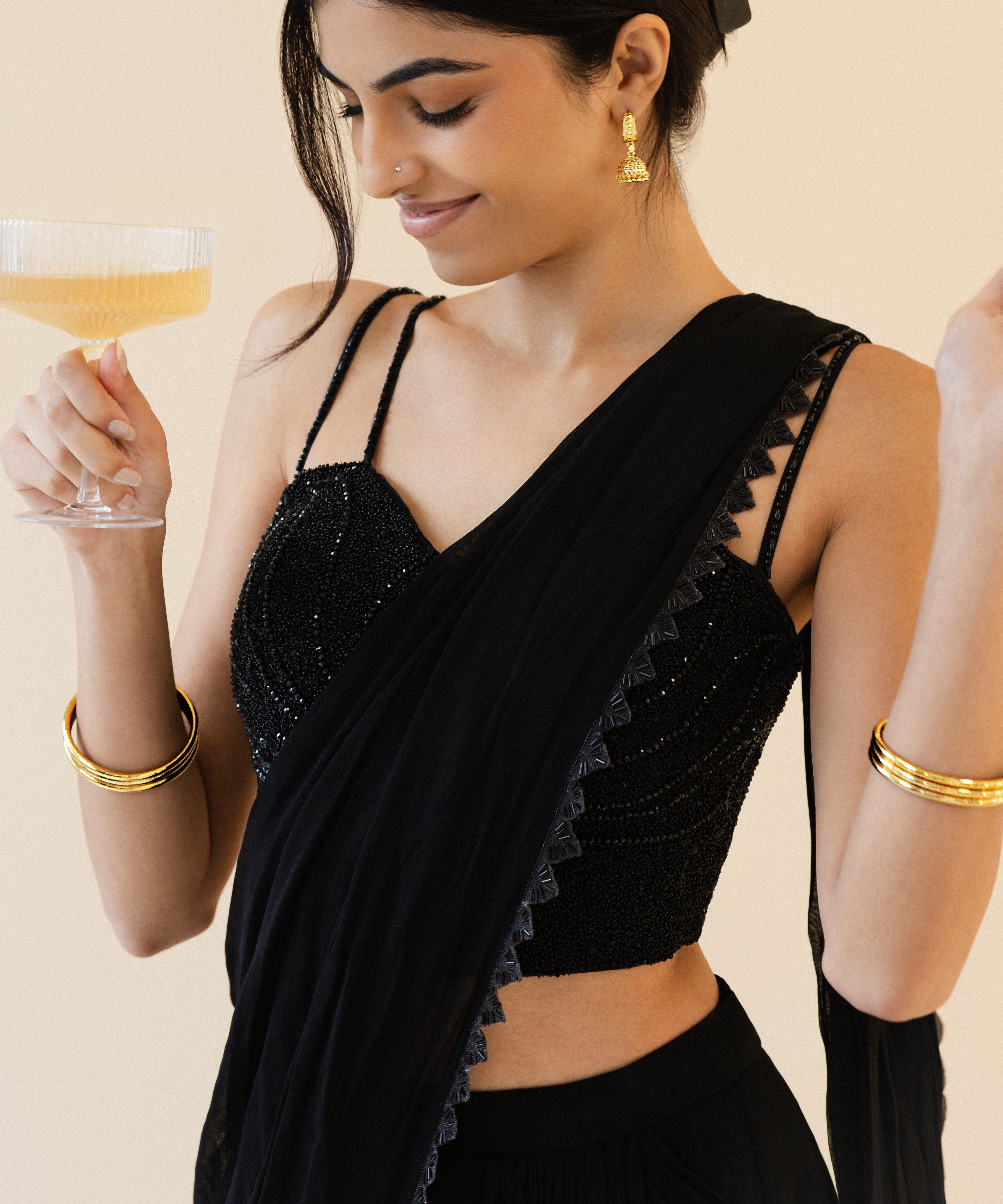 Woman in a black saree holding a glass of champagne against a beige background