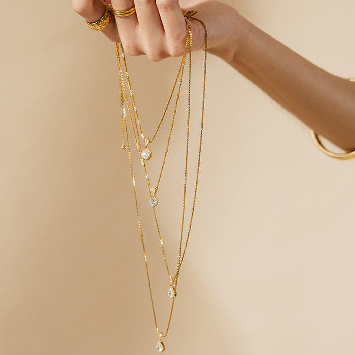 Hand holding multiple gold necklaces with small pendants against a beige background