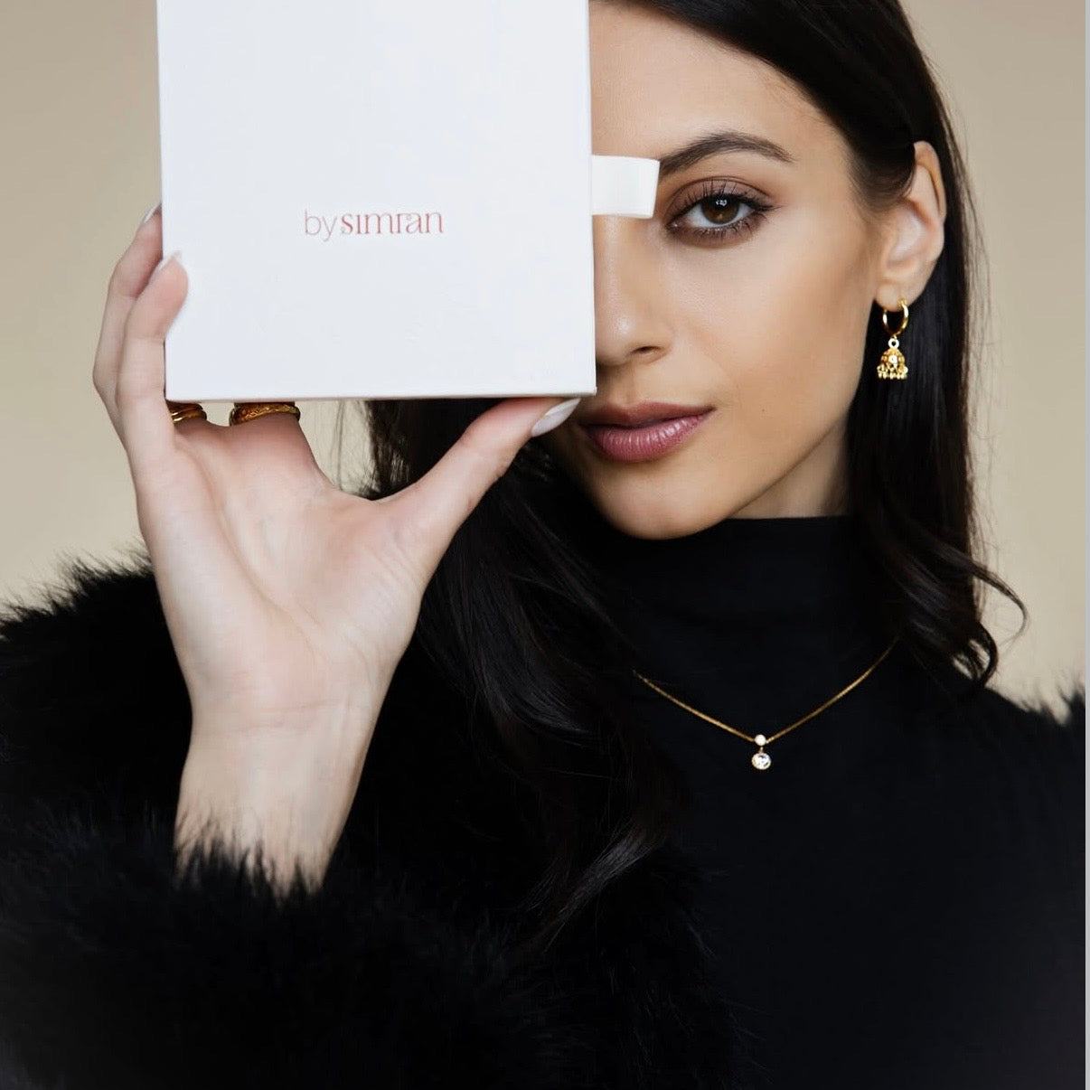 Woman holding a white box with 'by simran' branding, wearing gold jewelry.
